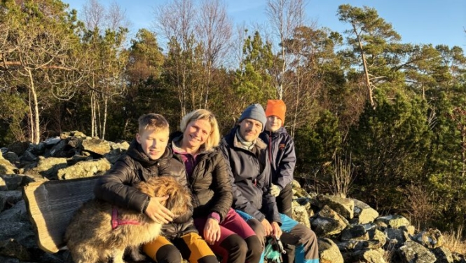 Švānu ģimene (tētis, mamma un divi dēli) ar suni sēž uz soliņa ar akmeņiem fonā.