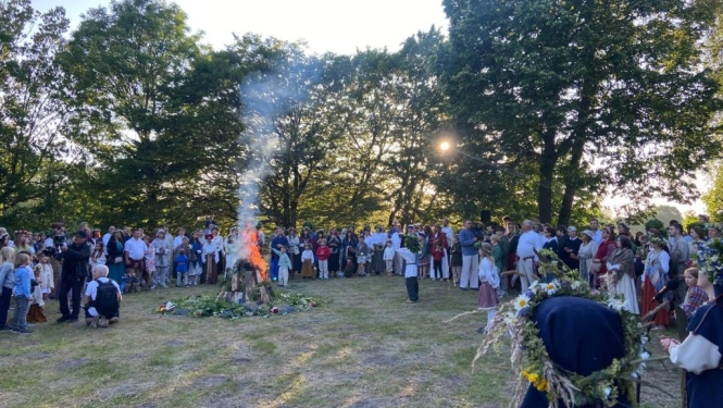 Dalībnieki 3×3 pasaules latviešu saietā Grobiņā tautastērpos ar vainagiem pulcējušies ap lielu ugunskuru.