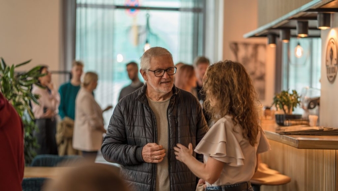 Vecāka gadagājuma vīrietis un jaunāka sieviete sarunājas pasākuma laikā kafejnīcas telpās, fonā redzami citi apmeklētāji.