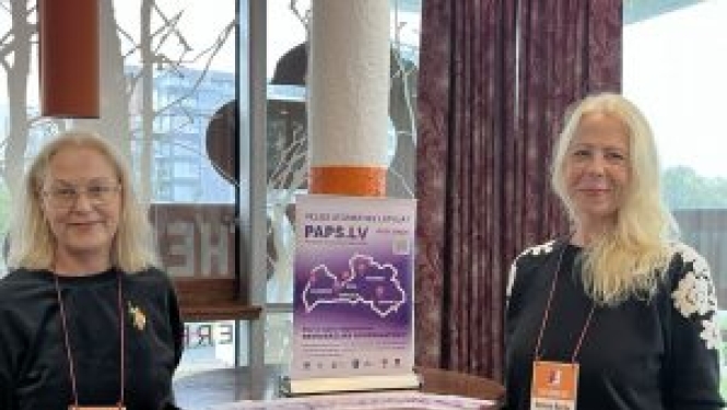 Rīgas reģiona remigrācijas koordinatore Daina Šulca un Kurzemes reģiona remigrācijas koordinatore Agnese Berģe stāv pie stāvgalda ar remigrācijas koordinatoru baneri pa vidu.