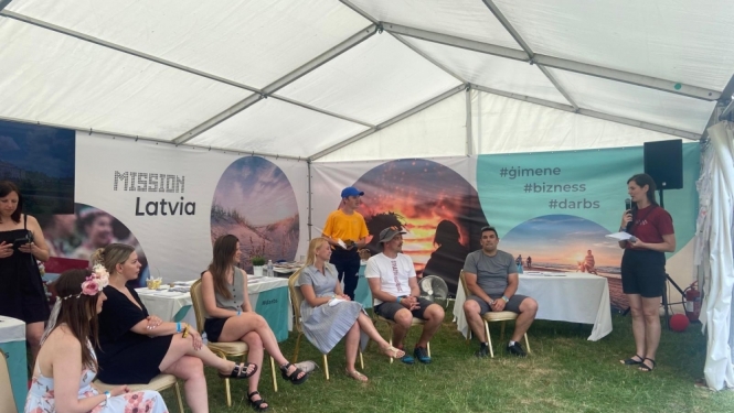 Vidzemes reģiona remigrācijas koordinatore Inga Madžule stāv informatīvajā teltī "Latvijas māja" Jāņu festivālā "Bērzes strazdi" pie galda, kur izvietoti bukleti, fonā redzami Latvijas attēli.