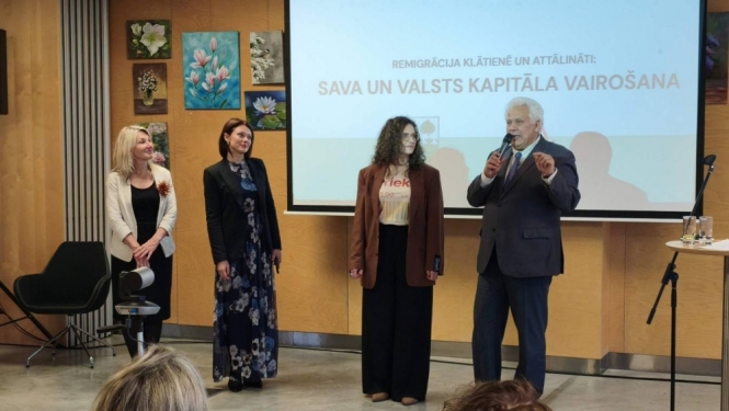 Pasākuma vadītājs Aigars Strauss pie ekrāna ar mikrofonu kopā ar trīs sievietēm - pasākuma organizatorēm.