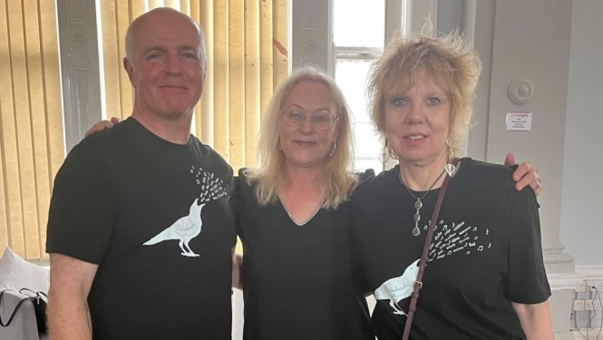 Sieviete formālā apģērbā stāv iekštelpās uz informatīva stenda fona ar ainavām, rokās turot sarkanu un dzeltenu rožu pušķi