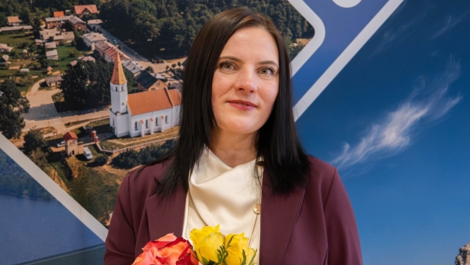 Sieviete formālā apģērbā stāv iekštelpās uz informatīva stenda fona ar ainavām, rokās turot sarkanu un dzeltenu rožu pušķi