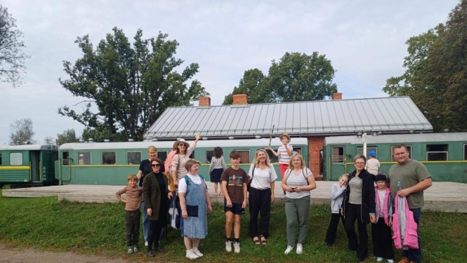 Dalībnieki remigrantu tīklošanās dienā “Manas mājas Vidzemē” uz bānīša fona pie Stāmerienas dzelzceļa stacijas.