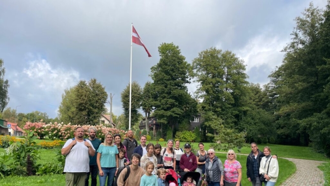 Dažāda vecuma remigrantu kopbilde pasākumā Vidzemē ar kokiem, ziediem un Latvijas karoga mastu fonā.