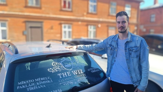 Uzņēmējs Saimons Burvis stāv pie sava auto Valmierā ar oranžu koka ēku fonā.