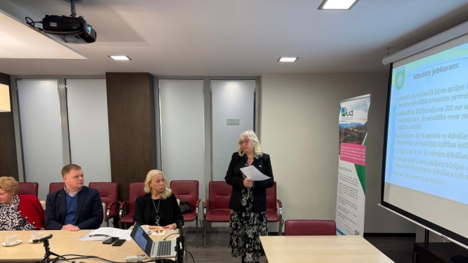 Sieviete stāv pie prezentācijas ekrāna un redzami trīs klausītāji.