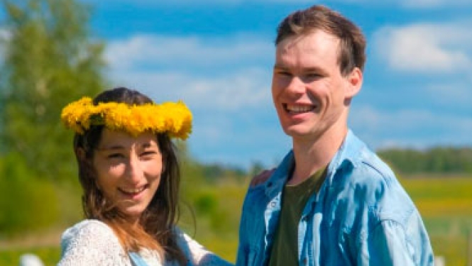 Jauns pāris smaida ziedošā pļavā Kurzemes laukos; sievietei galvā dzeltenu ziedu vainags