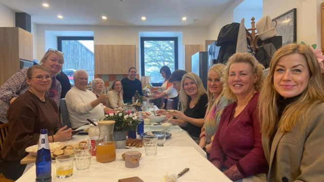 Cilvēku grupa sēž pie gara galda gaišā telpā un smaida kamerai.