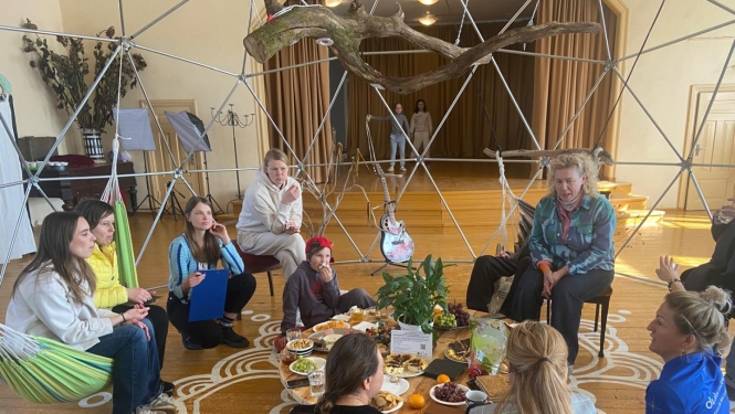 Cilvēku grupa sēž aplī uz koka grīdas gaišā, radošā telpā Augstūjā, Vidzemē, kopīgā pasākumā “Ciemos pie atbraucējiem"