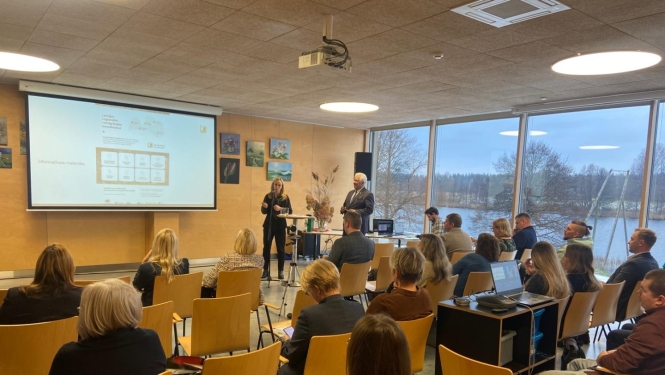 Konferenču zālē notiek prezentācija: pie ekrāna runātāji, zālē sēž klausītāji, fonā lieli logi ar skatu uz dabu