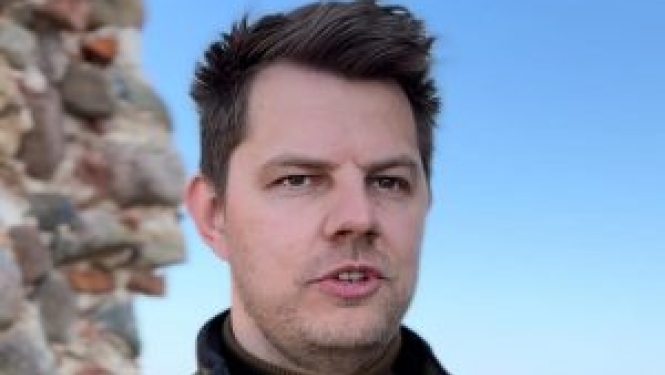 Vīrietis ar īsiem tumšiem matiem stāv ārā pie akmens mūra, fonā redzamas debesis