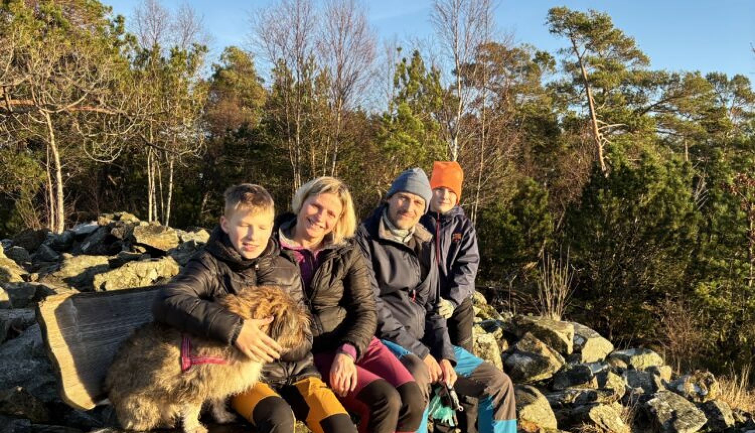 Švānu ģimene (tētis, mamma un divi dēli) ar suni sēž uz soliņa ar akmeņiem fonā.