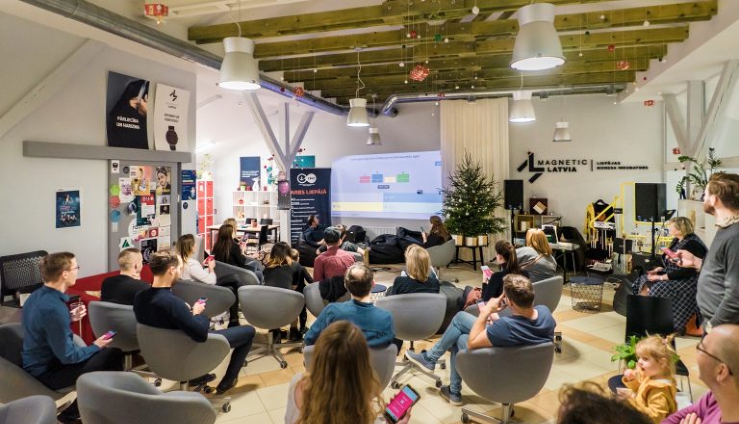 Mūsdienīga pasākumu telpa, kur sēž cilvēki un vēro prezentāciju ekrānā.