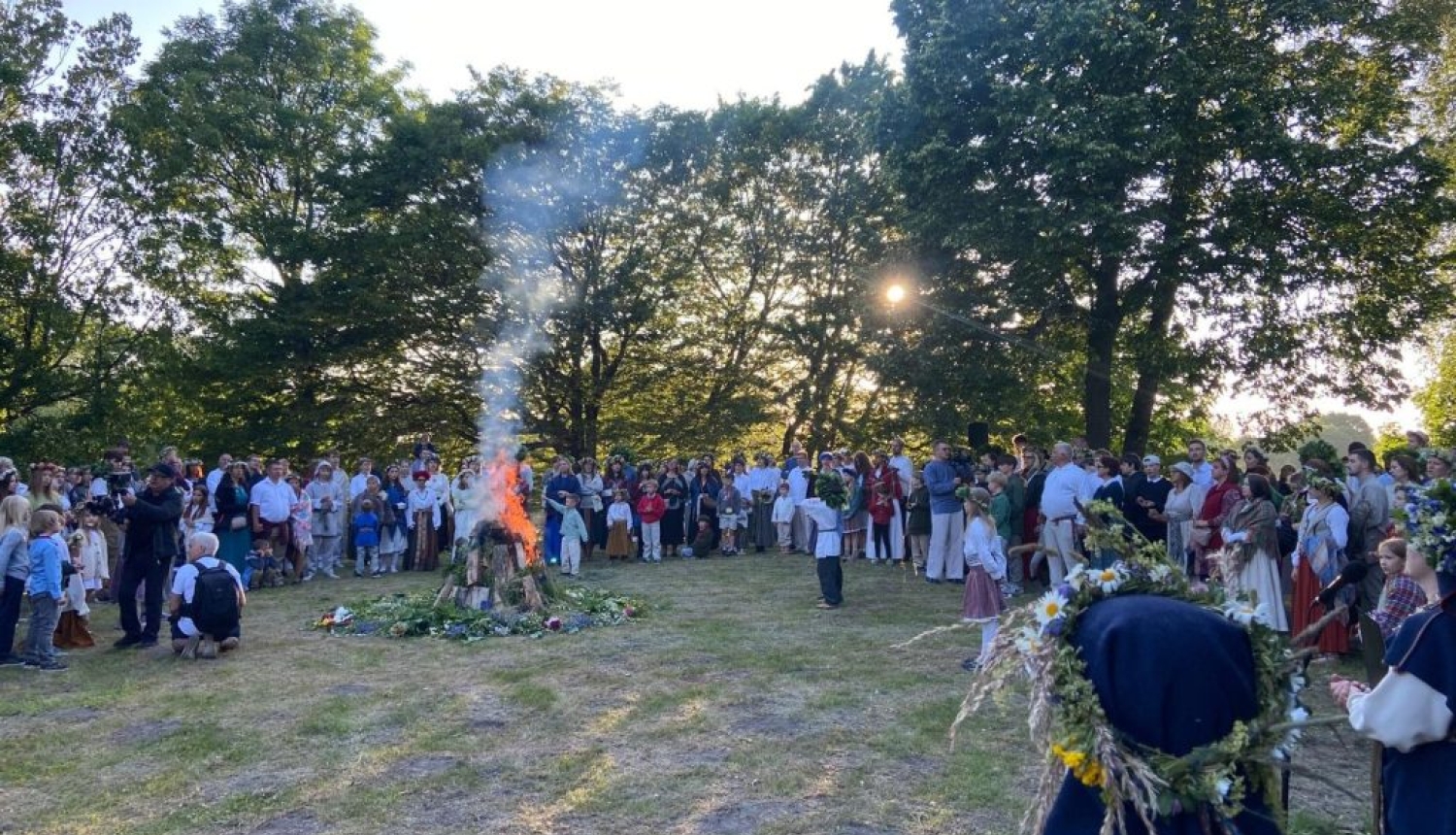 Dalībnieki 3×3 pasaules latviešu saietā Grobiņā tautastērpos ar vainagiem pulcējušies ap lielu ugunskuru.