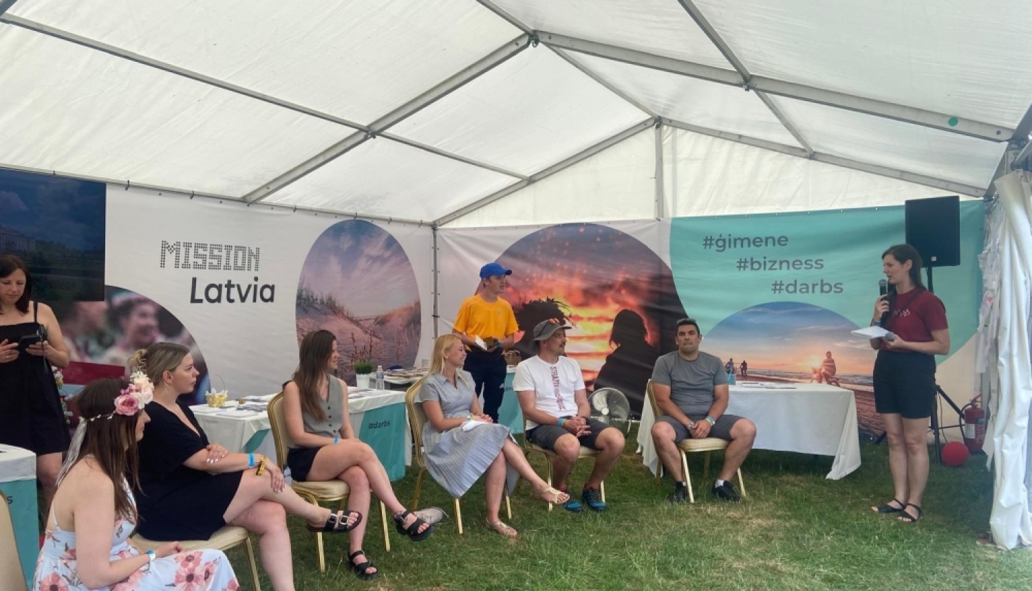 Vidzemes reģiona remigrācijas koordinatore Inga Madžule stāv informatīvajā teltī "Latvijas māja" Jāņu festivālā "Bērzes strazdi" pie galda, kur izvietoti bukleti, fonā redzami Latvijas attēli.