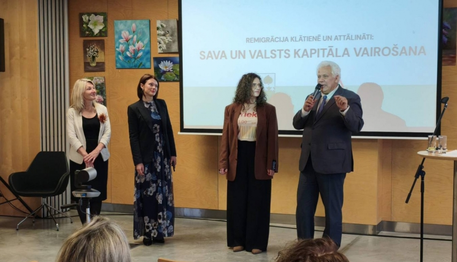 Pasākuma vadītājs Aigars Strauss pie ekrāna ar mikrofonu kopā ar trīs sievietēm - pasākuma organizatorēm.