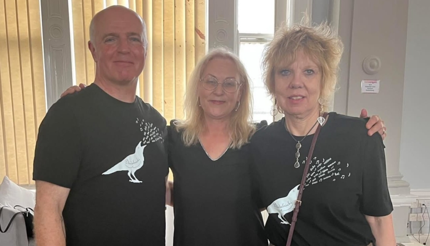 Sieviete formālā apģērbā stāv iekštelpās uz informatīva stenda fona ar ainavām, rokās turot sarkanu un dzeltenu rožu pušķi