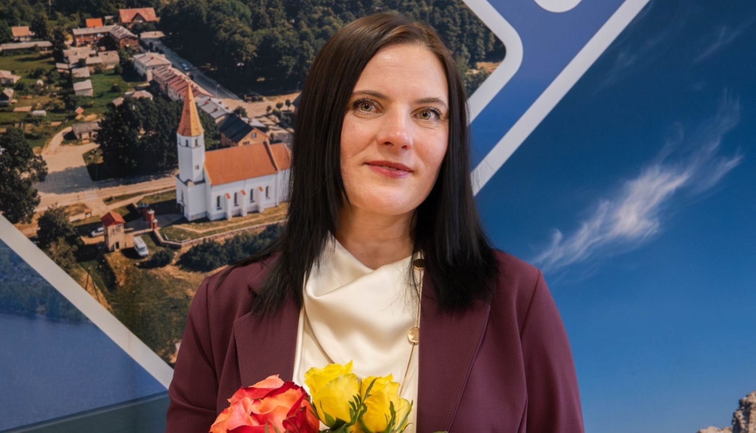 Sieviete formālā apģērbā stāv iekštelpās uz informatīva stenda fona ar ainavām, rokās turot sarkanu un dzeltenu rožu pušķi