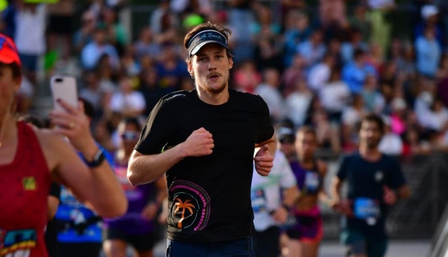 Edgars Prūsis skrien maratonā Maiami, finiša posmā starp citiem skrējējiem, simbolizējot aktīvu dzīvesveidu un atgriešanās ceļu mājās..