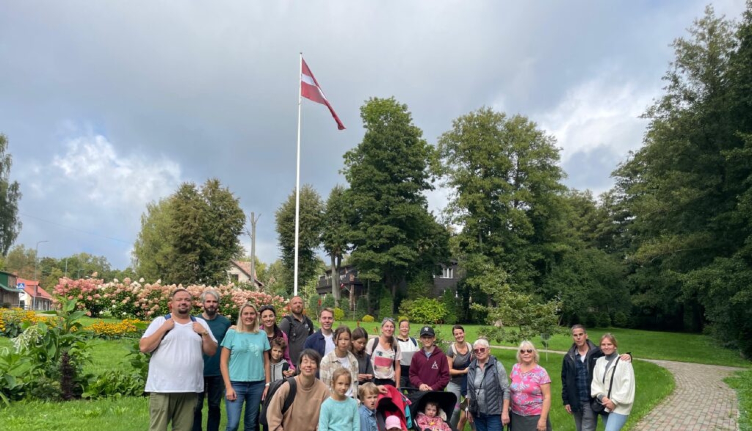 Dažāda vecuma remigrantu kopbilde pasākumā Vidzemē ar kokiem, ziediem un Latvijas karoga mastu fonā.