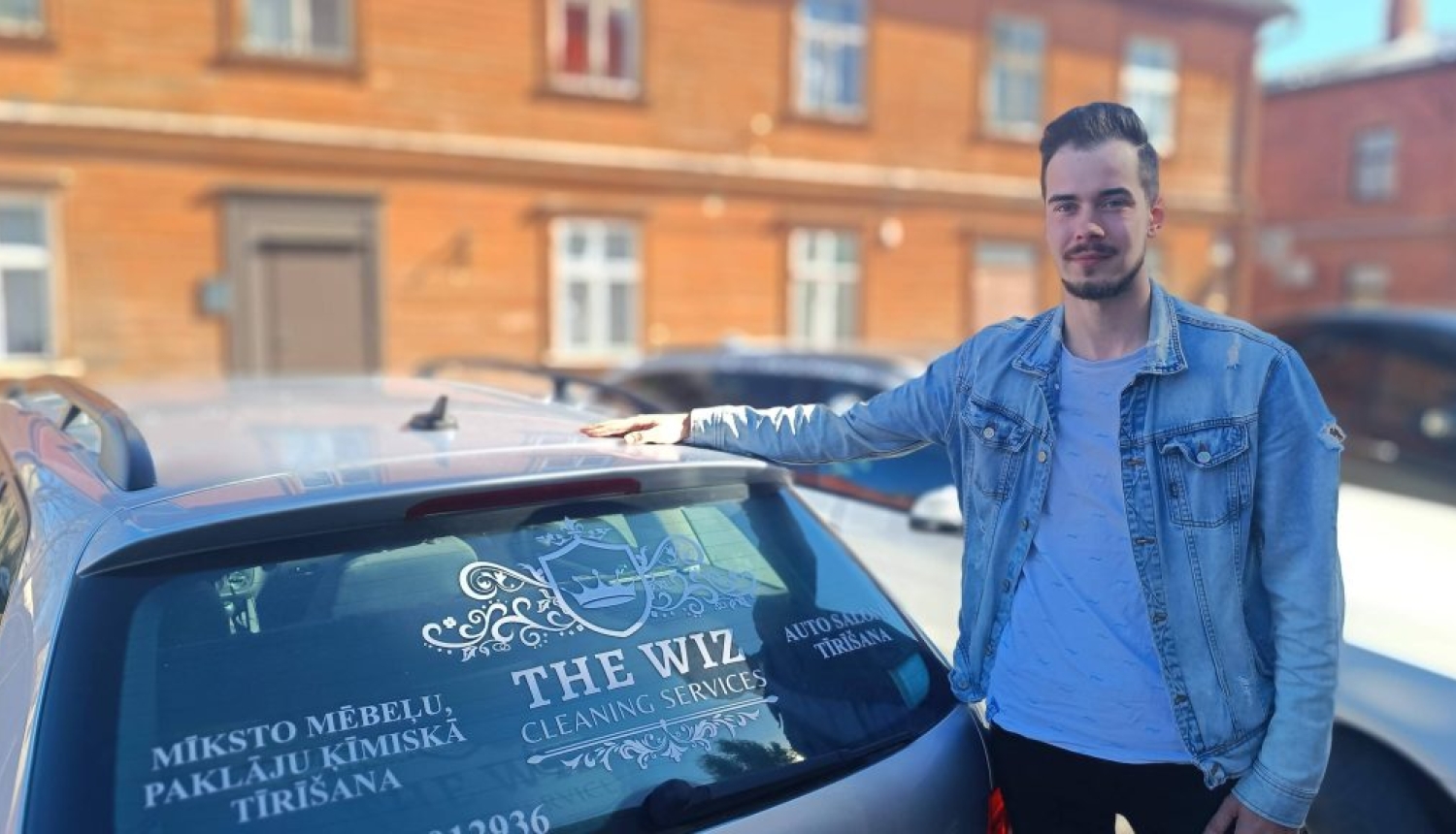 Uzņēmējs Saimons Burvis stāv pie sava auto Valmierā ar oranžu koka ēku fonā.