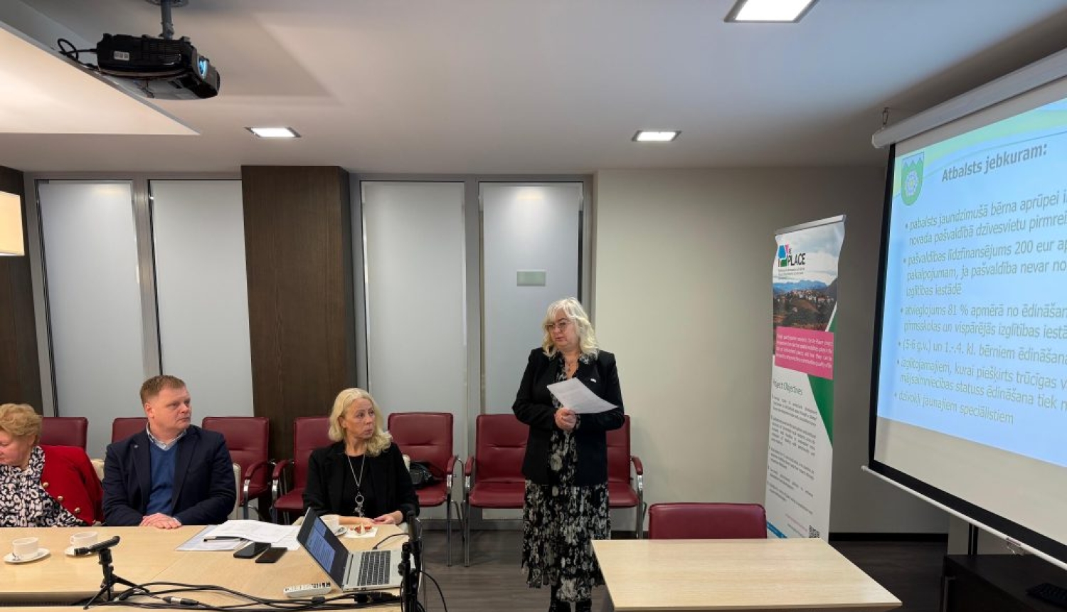 Sieviete stāv pie prezentācijas ekrāna un redzami trīs klausītāji.