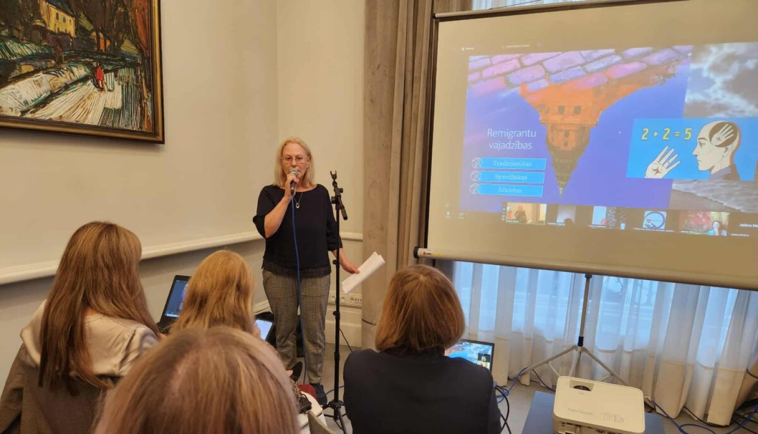 Rīgas reģiona remigrācijas koordinatore Daina Šulca stāv pie ekrāna un rāda prezentāciju klausītāju auditorijai.