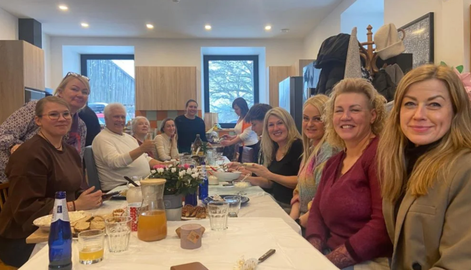 Cilvēku grupa sēž pie gara galda gaišā telpā un smaida kamerai.