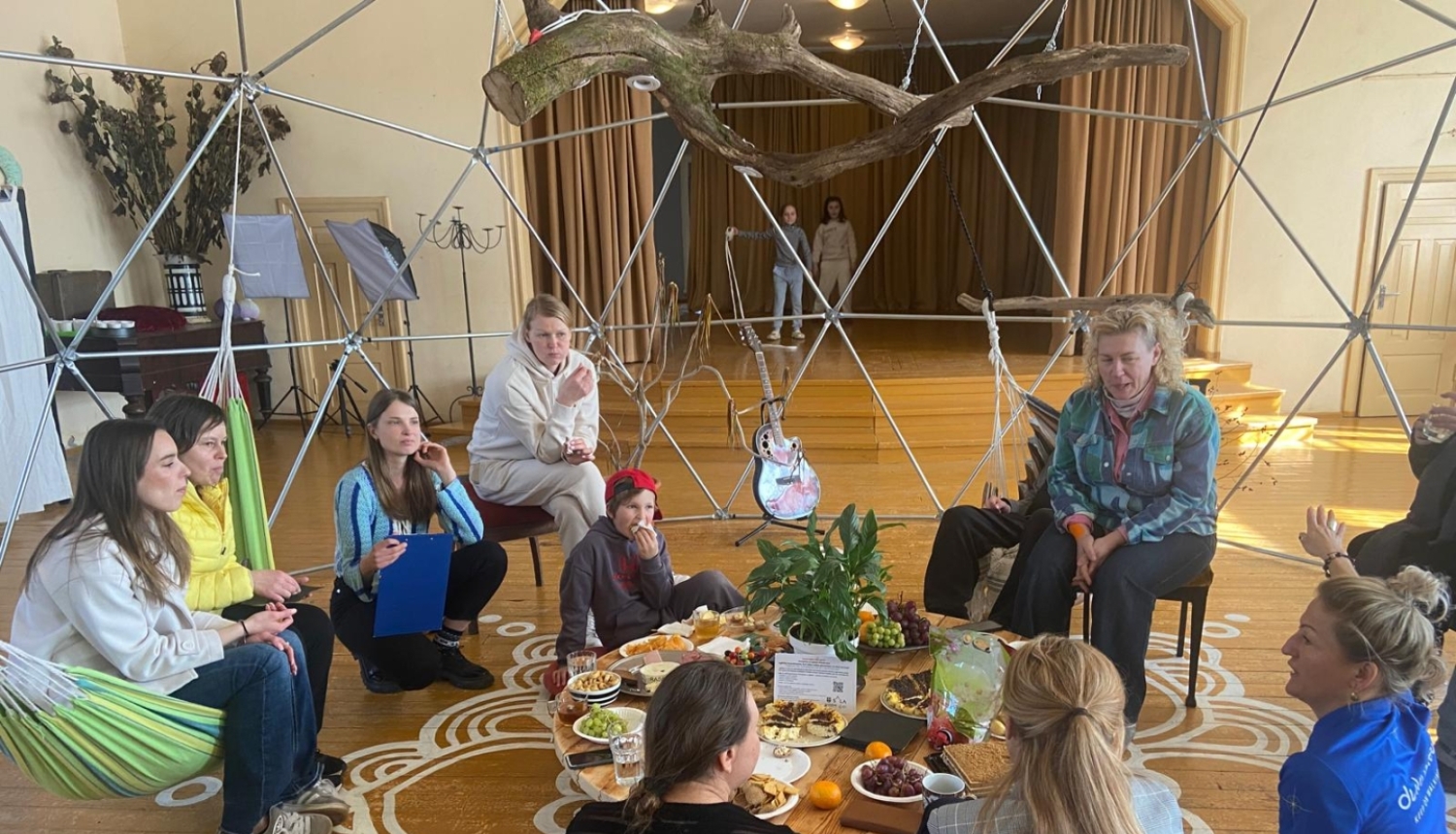 Cilvēku grupa sēž aplī uz koka grīdas gaišā, radošā telpā Augstūjā, Vidzemē, kopīgā pasākumā “Ciemos pie atbraucējiem"