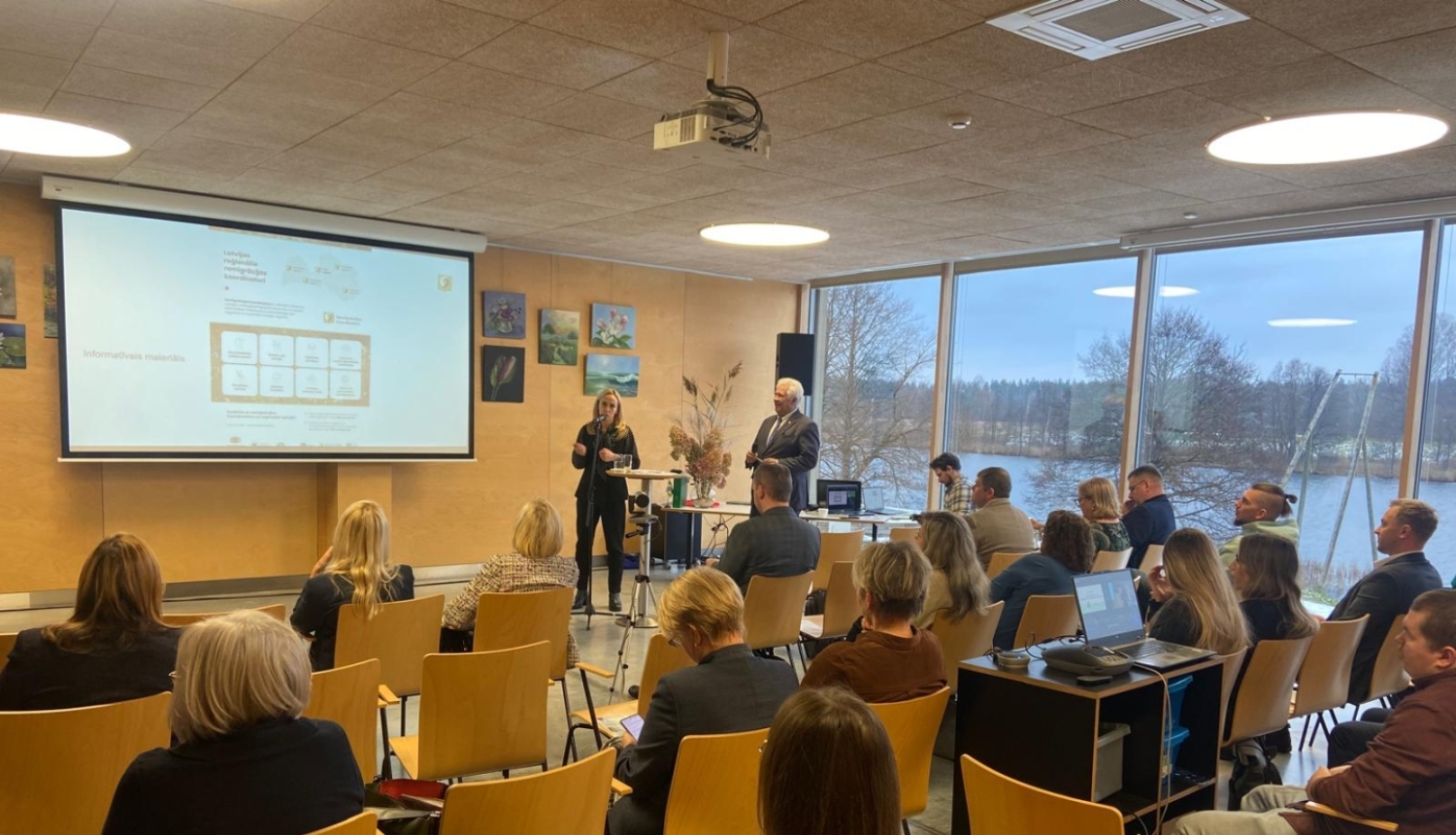 Konferenču zālē notiek prezentācija: pie ekrāna runātāji, zālē sēž klausītāji, fonā lieli logi ar skatu uz dabu