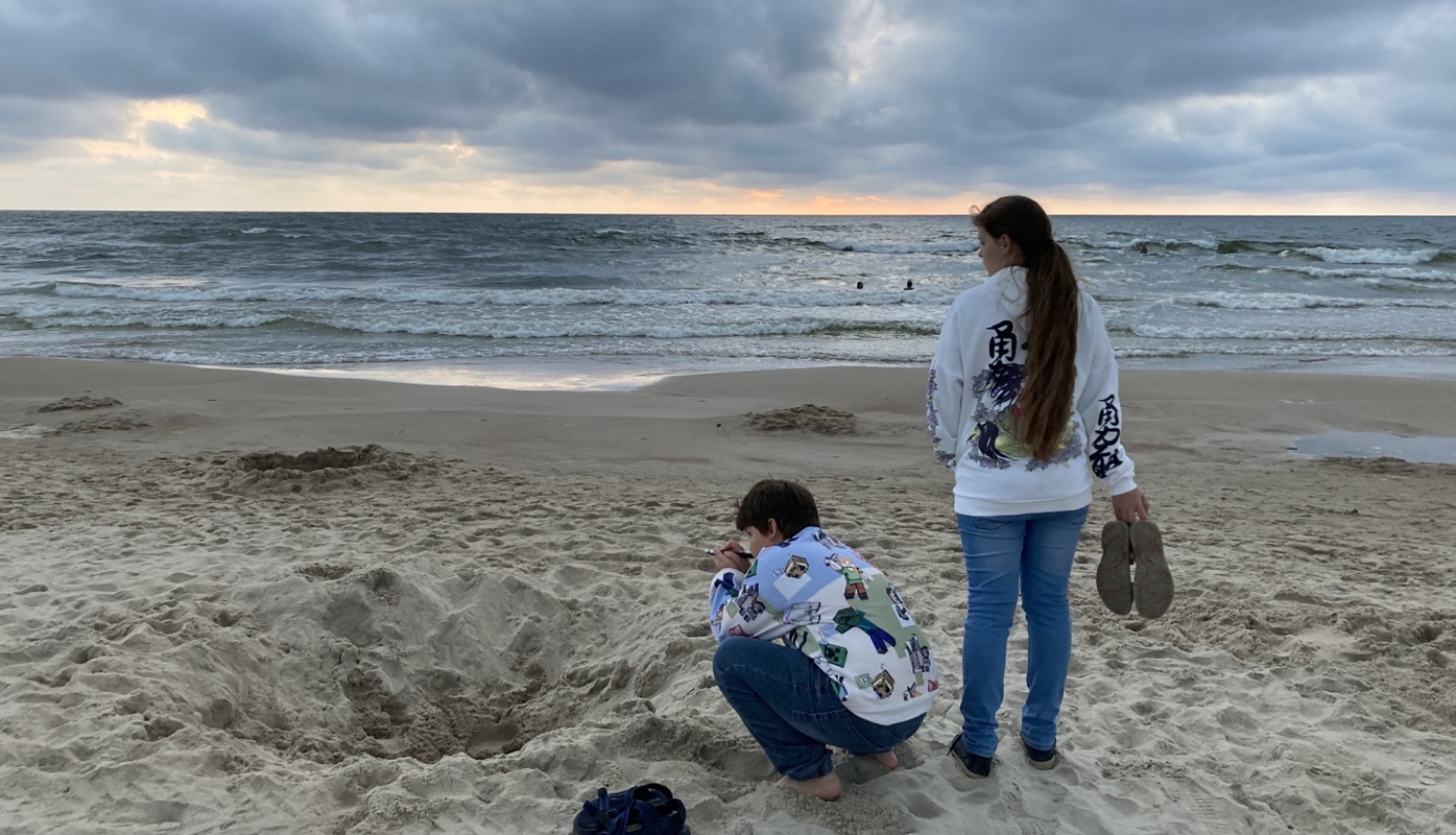 Meitene un zēns atrodas jūras krastā pievakarē, zēns spēlējas ar smiltīm, bet meitene raugās tālumā