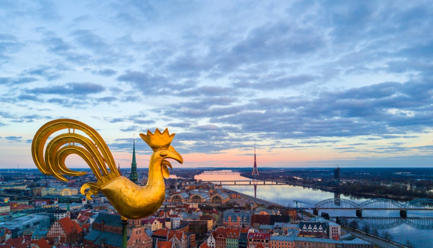 Rīgas panorāmas skats, kur attēlots baznīcas zelta gailis un teksts “Atgriezies Rīgā un reģionā: Rīga, Jūrmala + Ādažu, Salaspils, Siguldas, Mārupes, Olaines, Ķekavas, Ropažu novads".