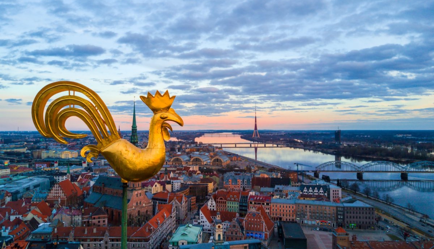 Rīgas panorāmas skats, kurā attēlots baznīcas zelta gailis un teksts “Atgriezies Rīgā un reģionā: Rīga, Jūrmala + Ādažu, Salaspils, Siguldas, Mārupes, Olaines, Ķekavas, Ropažu novads".